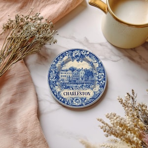 May include: Round blue and white Charleston coaster with a floral border. The coaster features a detailed illustration of Charleston buildings and the city's name in a banner. A mug of coffee sits nearby.