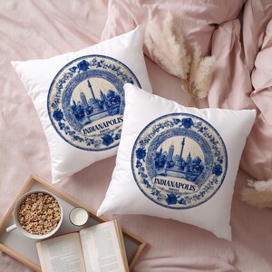 May include: Two white throw pillows with a blue and cream design featuring the Indianapolis skyline and the city's name. The pillows are on a pink bedspread, with a tray of cereal and a book in the foreground.