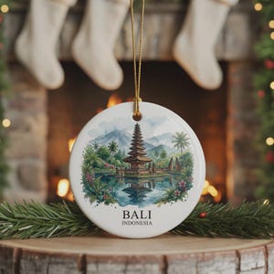 Könnte beinhalten: Ein rundes, weißes Keramikornament mit einer Aquarellillustration eines balinesischen Tempels an einem See mit Bergen im Hintergrund. Der Text BALI INDONESIA ist unter der Illustration in Schwarz aufgedruckt. Es hängt an einer goldenen Schnur.