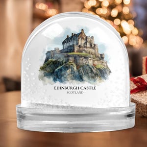 May include: A clear glass snow globe featuring a watercolor painting of Edinburgh Castle in Scotland. The castle is depicted on a rocky hill with a blue sky background. The text "EDINBURGH CASTLE SCOTLAND" is printed below the image.