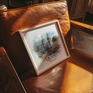 May include: A framed watercolour print of Quito, Ecuador, showing a cathedral with two spired towers. The artwork is in a light wooden frame, resting on a brown leather armchair. The print includes the city's name.