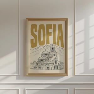 May include: A framed wall art print featuring the word SOFIA in large gold letters above a black line drawing of the Alexander Nevsky Cathedral in Bulgaria, set against a light cream background with a mountain range outline behind the building.