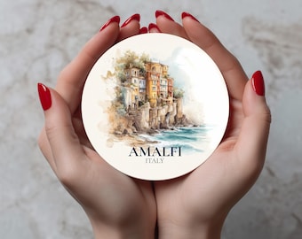 May include: A round coaster featuring a watercolor painting of Amalfi, Italy. The artwork depicts colorful buildings on a cliffside overlooking the sea. The text "AMALFI ITALY" is printed at the bottom.