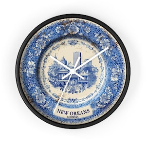 May include: Round wall clock with a black frame and a blue and white floral design. The clock face features a New Orleans cityscape illustration and the text "NEW OREANS LOUISIANA". The clock hands are white.