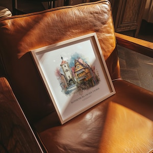 May include: A framed watercolour print of Rothenburg ob der Tauber, Germany, showing colourful buildings and a tower. The print has a light wooden frame and the text "ROTHENBURG OB DER TAUBER GERMANY". It rests on a brown leather chair.