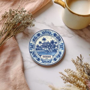 May include: Round blue and white ceramic coaster featuring a detailed illustration of a Japanese building and palm trees. The coaster has the word "OKINAWA JAPAN" printed on it. The coaster is surrounded by a floral design.