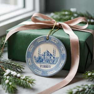 Peut inclure: Ornement rond en céramique bleu et blanc représentant une mosquée avec des minarets, avec l'inscription "TURKEY". L'ornement est suspendu devant un coffret cadeau vert emballé avec un ruban rose.
