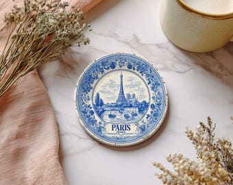 May include: Round blue and white ceramic coaster featuring the Eiffel Tower and the word "PARIS". The coaster has a floral border design and is surrounded by dried flowers and a mug of coffee.