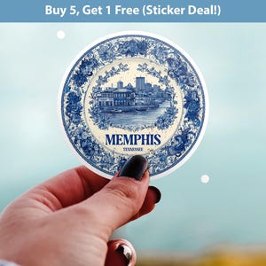 May include: A round sticker featuring a blue and white illustration of Memphis, Tennessee. The design includes a cityscape, floral border, and the city's name. The text "Buy 5, Get 1 Free (Sticker Deal!)" is at the top.