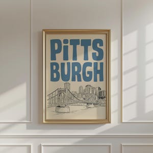 Könnte beinhalten: Ein gerahmter Kunstdruck mit dem Wort PITTSBURGH in großen, fetten, blauen serifenlosen Buchstaben über einer detaillierten schwarz-weißen Strichzeichnung einer Hängebrücke und der Skyline der Stadt auf hellcremefarbenem Hintergrund.