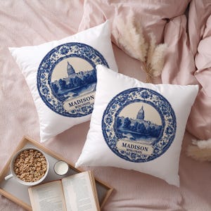 May include: Two white square throw pillows with a blue and white design featuring the Madison, Wisconsin capitol building. The pillows have a floral border and the city name. The pillows are on a pink bed.