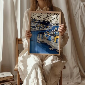 K&ouml;nnte beinhalten: Ein gerahmter Kunstdruck, der eine Stadtlandschaft mit blauem Wasser und Geb&auml;uden in Blau-, Gelb- und Wei&szlig;t&ouml;nen zeigt. Das Kunstwerk hat oben ein gepunktetes Muster und ist in einem hellen Holzrahmen gehalten.