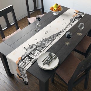 Könnte beinhalten: Cremefarbener Tischläufer mit einem schwarz-weißen Stadtbilddesign. Der Läufer zeigt eine detaillierte Illustration von Gebäuden, Bäumen und einer Brücke. Akzentuiert mit pfirsichfarbenen Wolken, liegt er auf einem dunklen Holztisch mit Gedecken.