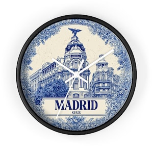 May include: Round wall clock with a black frame featuring a blue and white illustration of a building in Madrid, Spain. The clock face includes the word "MADRID" and floral accents.