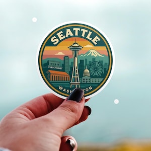 May include: A round sticker featuring a Seattle, Washington design. The sticker includes the Space Needle, mountains, and the text "SEATTLE WASHINGTON". The design is in shades of green, orange, and blue.