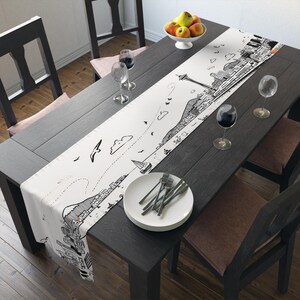 Könnte beinhalten: Weißer Tischläufer mit einem schwarz-weißen, illustrierten Stadtlandschaftsdesign. Der Läufer zeigt eine detaillierte Zeichnung einer Skyline, Segelbooten und Vögeln. Er liegt auf einem dunklen Holztisch, der für ein Essen gedeckt ist.