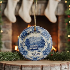May include: Round ceramic Christmas ornament with a blue and white floral design. The center features a detailed illustration of a Lisbon, Portugal, street scene with a tram. The ornament is suspended by a gold string.
