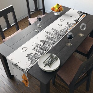 May include: A white table runner with a black and orange cityscape design. The runner features a detailed illustration of a city skyline, with buildings and bridges. The runner is placed on a dark wooden table, set for a meal with plates, silverware, and wine glasses.