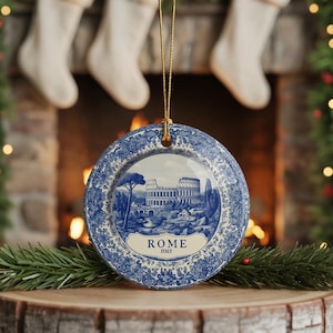 May include: A round, blue and white ceramic ornament featuring a detailed illustration of the Colosseum in Rome. The ornament has a floral border and a gold string for hanging. The word "ROME" is displayed on the ornament.