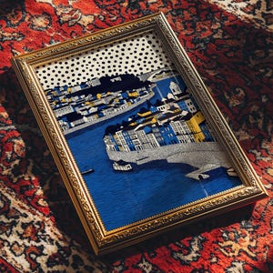 K&ouml;nnte beinhalten: Ein gerahmter Kunstdruck mit einer detaillierten Illustration einer Stadtlandschaft mit Fluss, Br&uuml;cke und Geb&auml;uden. Das Kunstwerk befindet sich in einem goldfarbenen Rahmen und liegt auf einem roten gemusterten Teppich.
