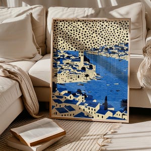 May include: A framed art print showing a coastal town with blue water and white buildings with blue roofs. The artwork has a beige background with black polka dots. The print is in a wooden frame and is leaning against a beige sofa.
