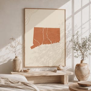 May include: A framed wall art print featuring a terracotta-colored silhouette of the state of Connecticut on a cream background. The map shape includes white line details and is accented by thin, curved line patterns in the top left and bottom right.