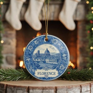 May include: A round, blue and white ceramic Christmas ornament featuring a detailed illustration of Florence, Italy. The ornament has a gold string for hanging and is decorated with floral patterns. The word "FLORENCE" is printed on the ornament.