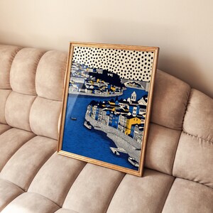 K&ouml;nnte beinhalten: Ein gerahmter Kunstdruck, der eine Stadtansicht mit blauem Wasser, Geb&auml;uden in Blau-, Gelb- und Wei&szlig;t&ouml;nen und einer Br&uuml;cke zeigt. Das Kunstwerk hat einen goldenen Rahmen und einen gepunkteten schwarz-wei&szlig;en Himmel. Der Druck wird auf einem beigen Sofa pr&auml;sentiert.