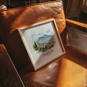 May include: A framed watercolor print of Naples, Italy, featuring a scenic view of a coastal town with mountains in the background. The print has a light wooden frame and the text "NAPLES ITALY" at the bottom.