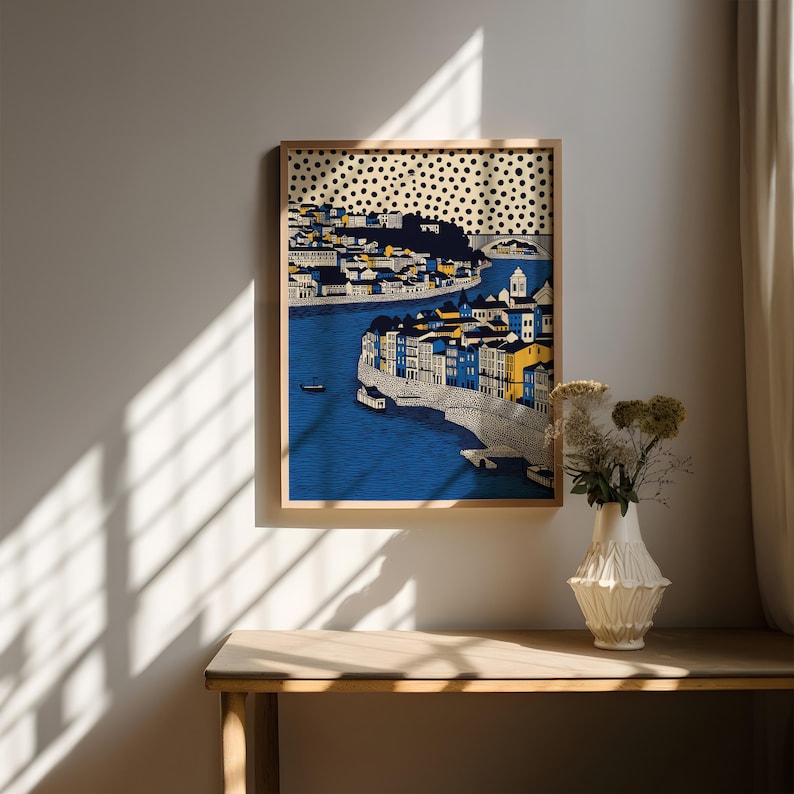 K&ouml;nnte beinhalten: Ein gerahmter Kunstdruck, der eine K&uuml;stenstadt mit blauem Wasser und Geb&auml;uden in Blau, Gelb und Wei&szlig; zeigt. Das Kunstwerk hat oben ein gepunktetes Muster und wird an einer Wand &uuml;ber einem Holztisch ausgestellt.