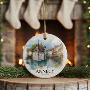 May include: A round ceramic Christmas ornament featuring a watercolor painting of Annecy, France. The image shows a building on a canal with surrounding buildings. The text "Annecy France" is printed below the image. The ornament hangs from a gold string.