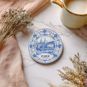 May include: A round, white coaster with a blue and white floral border and a central image of Zurich, Switzerland. The word "ZURICH" is printed in blue. A gold-rimmed mug with coffee sits nearby.