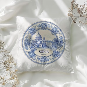 May include: A white square cushion with a blue and white decorative plate design. The plate shows a building with domes and the text "SOFIA BULGARIA" in a floral border. The cushion is on a white surface.