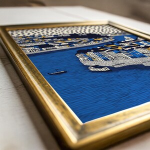 K&ouml;nnte beinhalten: Gerahmter Kunstdruck mit einer stilisierten Illustration einer K&uuml;stenstadt. Das Kunstwerk zeigt einen blauen Fluss, Geb&auml;ude in Wei&szlig;, Blau und Gelb sowie ein kleines Boot. Der goldfarbene Rahmen verleiht dem Bild eine elegante Note.