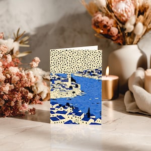 May include: A folded greeting card featuring a blue and white illustration of a coastal town with a tower, set against a blue sea. The top portion of the card has a cream background with black polka dots. The card is displayed on a marble surface.