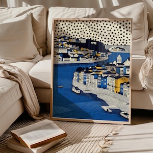 K&ouml;nnte beinhalten: Gerahmter Kunstdruck mit einer farbenfrohen Stadtlandschaft mit Fluss. Die Geb&auml;ude sind in Blau, Gelb und Wei&szlig; gehalten, mit einem gepunkteten Himmel. Der Druck wird auf einem beigen Sofa pr&auml;sentiert.