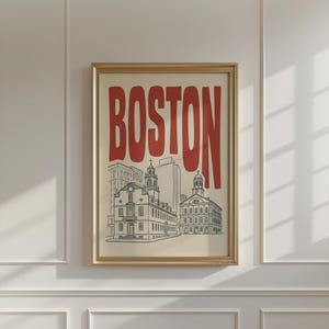 May include: A framed wall art print featuring the word BOSTON in large, bold red letters above a black and white line drawing of historic city buildings, including the Old State House and Faneuil Hall, set against a light cream background.