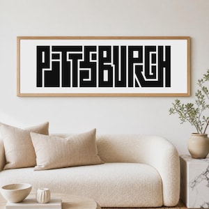 Peut inclure: Une œuvre d'art murale rectangulaire présentant le mot PITTSBURGH dans une police de caractères géométrique et épaisse de couleur noire sur fond blanc, encadrée par un cadre en bois clair. La typographie présente des lignes angulaires.