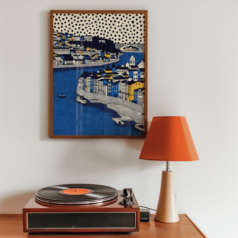 K&ouml;nnte beinhalten: Ein gerahmter Kunstdruck, der eine K&uuml;stenstadt mit blauem Wasser und farbenfrohen Geb&auml;uden zeigt. Ein Vintage-Plattenspieler mit einer Schallplatte und einem orangefarbenen Etikett befindet sich unter dem Kunstwerk. Ein orangefarbener Lampenschirm sitzt auf einem beigen Lampenfu&szlig;.