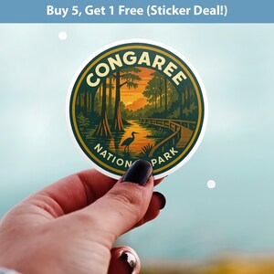 May include: A round sticker featuring the Congaree National Park logo. The design includes a scenic view of a river, trees, and a boardwalk, with a heron in the foreground. The sticker has a green and gold border and the text "CONGAREE NATIONAL PARK".