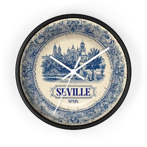 May include: Round wall clock with a black frame and a cream-colored face. The clock face features a blue and white illustration of a building and the text "SEVILLE SPAIN". The clock hands are white.