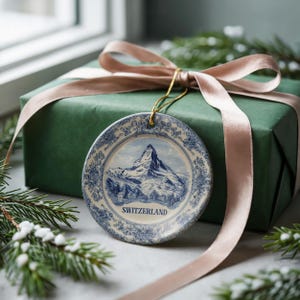 May include: A round ceramic ornament with a blue and white mountain scene and the word "SWITZERLAND". The ornament hangs in front of a wrapped green gift box with a pink ribbon. Decorative evergreen branches are in the foreground.