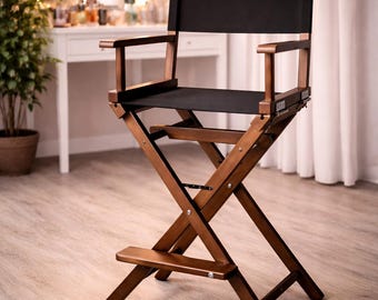 May include: A tall, brown wooden director's chair with a black canvas seat and back. The chair has armrests and a footrest. The background includes a vanity with a lighted mirror and a vase of flowers, suggesting a makeup or dressing area.