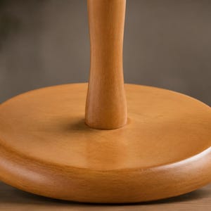 May include: A light brown wooden stand featuring a smooth, circular base and a central vertical post. The object has a natural wood grain finish with a polished surface, showcasing a simple, functional design with a rounded edge on the base.