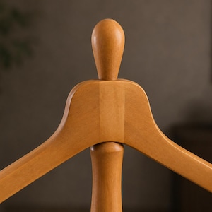 May include: A light brown wooden clothes hanger featuring a smooth, curved shoulder design and a central vertical post topped with a rounded, finial-like wooden knob. The hanger has a polished finish and a sturdy, solid construction.