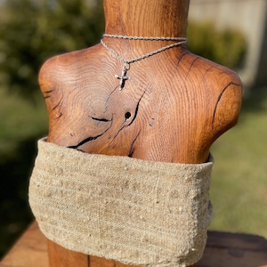 Könnte beinhalten: Eine silberne Kreuz-Anhänger-Halskette, die auf einer Holzbüste präsentiert wird. Die Büste besteht aus hellbraunem Holz und hat ein natürliches Maserungsmuster. Ein beiges Stoffband ist um die Büste gewickelt. Der Hintergrund ist verschwommen und zeigt grünes Gras und Bäume.