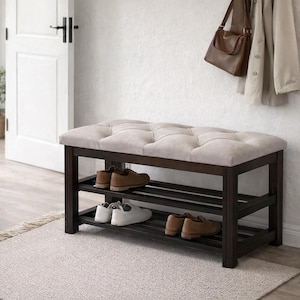 Può includere: Una panca scarpiera in legno marrone scuro con un cuscino di seduta beige trapuntato. La panca include due ripiani a doghe sottostanti, che ospitano attualmente due paia di scarpe in pelle marrone e un paio di scarpe da ginnastica bianche.