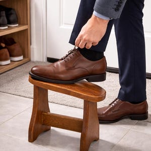 May include: A person tying the laces of a brown leather dress shoe. The shoe is on a small wooden stool. The person is wearing a navy blue suit and black socks. A shoe rack is visible in the background.