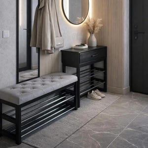 Könnte beinhalten: Moderne Eingangseinrichtung mit einer gesteppten grauen Bank mit schwarzem Metallrahmen und Schuhregal. Ein passender schwarzer Schrank mit Schublade und Schuhaufbewahrung steht daneben. Ein runder Spiegel und eine Garderobe vervollständigen den Raum.