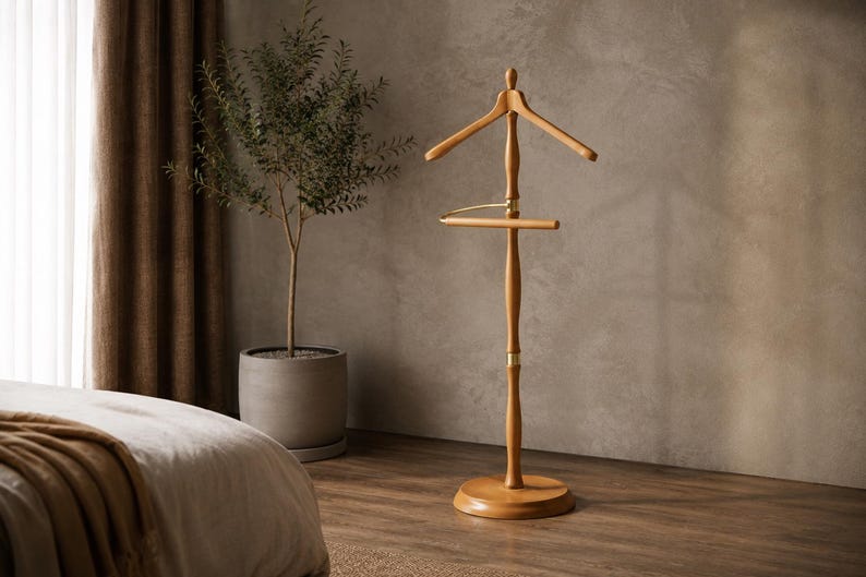 May include: A freestanding wooden valet stand featuring a smooth light brown finish, a contoured coat hanger top, a horizontal trouser bar, and a central vertical post supported by a circular base, accented with polished gold-tone metal hardware.